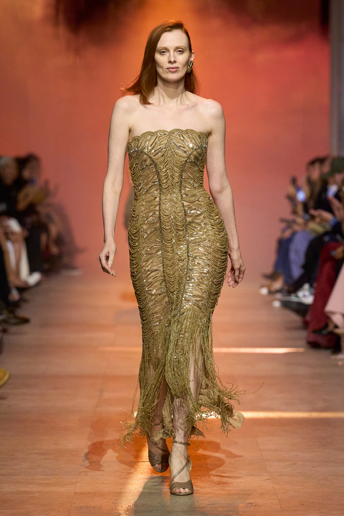 Model walking the runway in a strapless, gold beaded gown with intricate pattern and fringe at the hem, during a fashion show.
