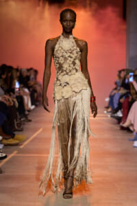 Model walking the runway in a beige lace halter gown with floral appliqué and long fringe skirt, on a warm orange backdrop.