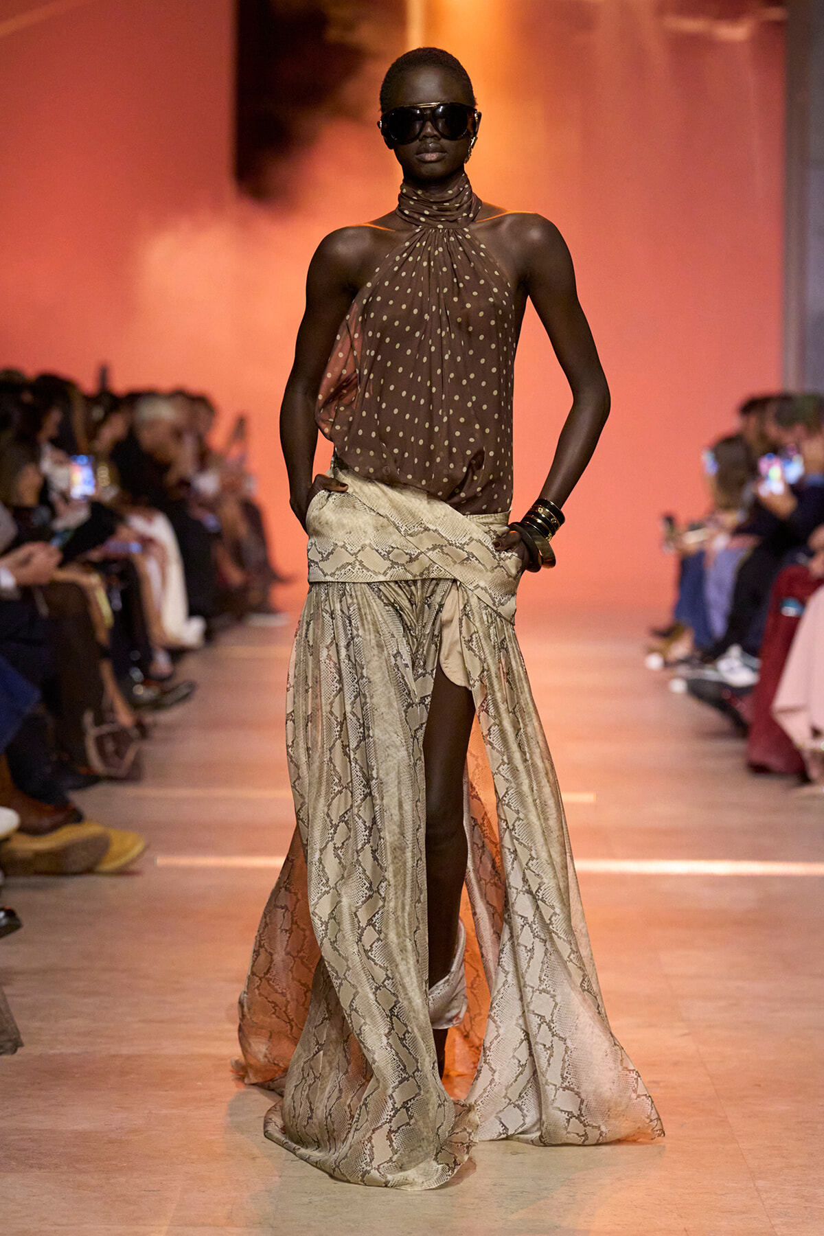 Model on a runway wearing a brown polka-dot halter top with flowing snakeskin-print pants, oversized sunglasses, and bracelets, orange backdrop.