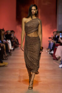 Model walks the runway in a sparkling one-shoulder brown draped two-piece gown with matching skirt and bangles, fashion show scene.