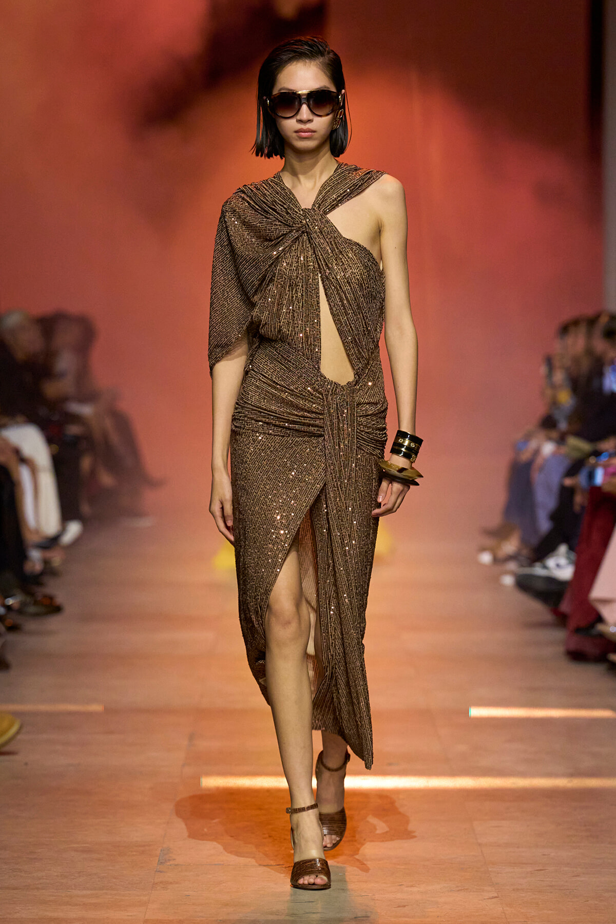 Model walks the runway in a brown sequined one-shoulder gown with a draped, cutout front and sunglasses.