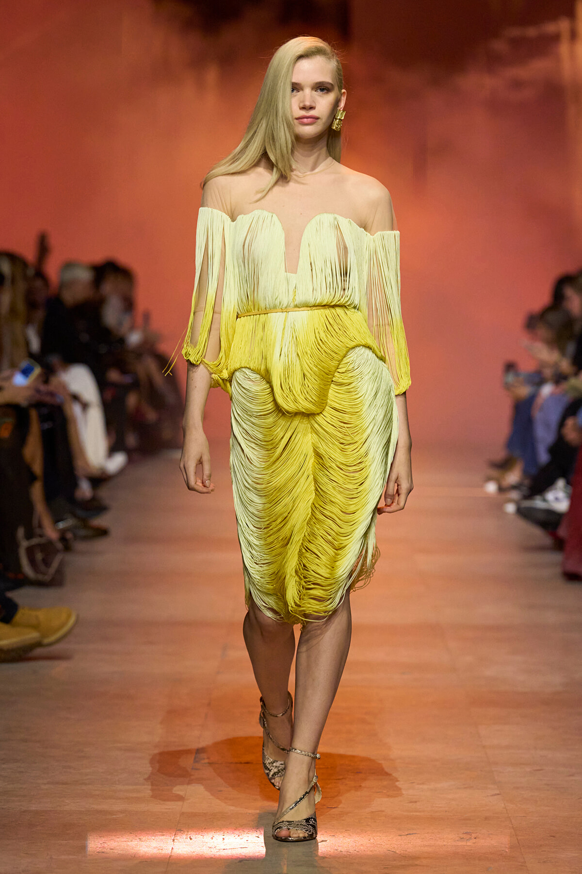 Model walking a runway in a yellow off-shoulder fringe dress with a deep V neckline and matching fringed skirt, audience blurred in the background.