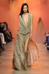 Model walks a fashion runway in a beige snakeskin–print, draped gown with a deep plunging V and high slit, orange backdrop behind her.