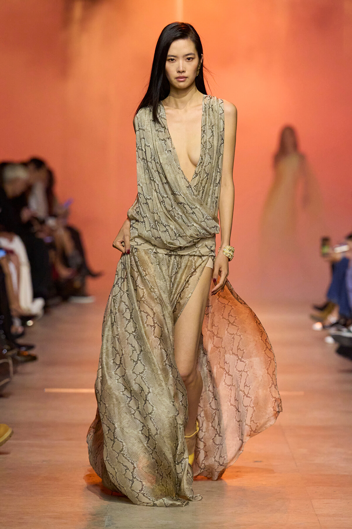 Model walks a fashion runway in a beige snakeskin–print, draped gown with a deep plunging V and high slit, orange backdrop behind her.
