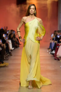 Model on a runway wearing a pale yellow, one-shoulder, flowing gown with draped pleats and gold accessories on the arm and ear.