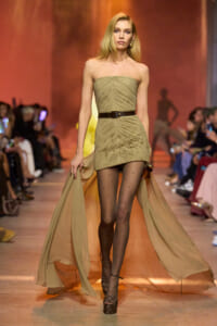Model on a runway wearing a strapless beige ruched mini dress with a brown belt and flowing sheer panels trailing behind her.