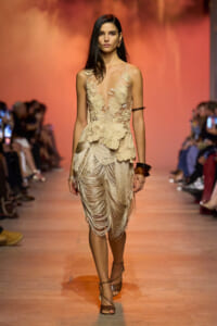 Model walking a runway in a sheer beige floral applique dress with a layered fringe skirt.