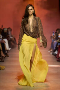 Model walks a runway in a brown polka-dot sheer blouse with deep drape paired with a bright yellow high-slit skirt.