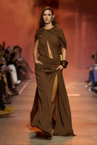 Model walking the runway in a brown draped gown with a thigh-high slit, hands in pockets, gold jewelry visible; warm stage lighting in background.