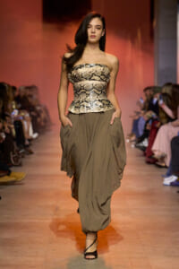 Model on a runway wearing a strapless snakeskin top and an olive draped skirt with pockets, walking confidently as the audience looks on.