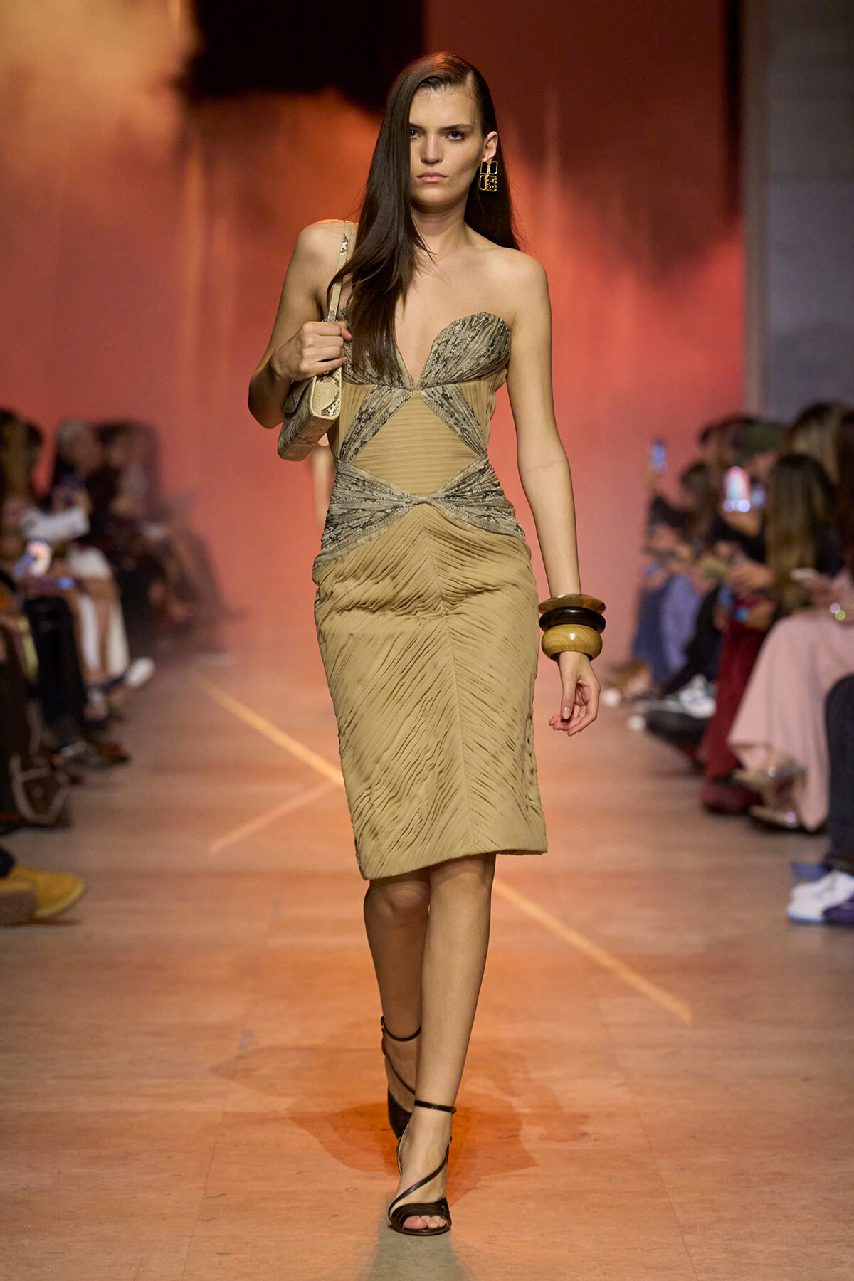 Model walks runway in a beige, ruched, form-fitting dress with metallic mesh accents and a plunging neckline. She accessorizes with large gold earrings, stacked bangles, and a small shoulder bag.