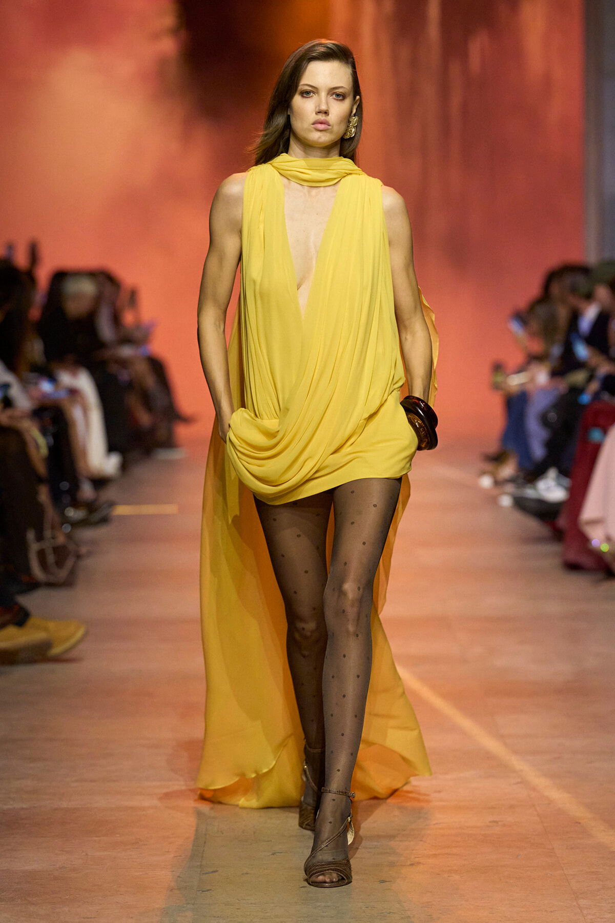 Model walks the runway in a bright yellow draped chiffon dress with a scarf-like neckline and cape-like back, paired with dotted tights and strappy sandals.