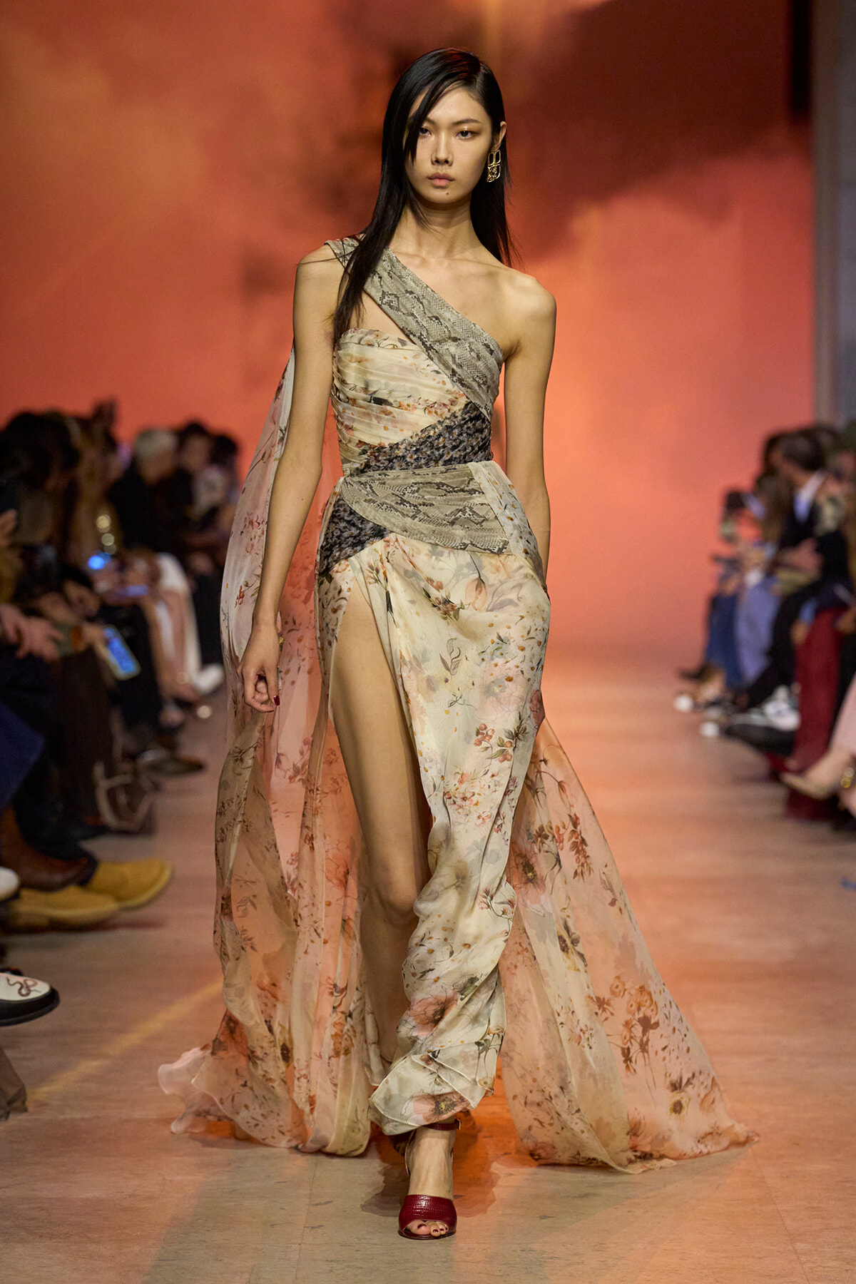 Model walks the runway in a pastel floral, one-shoulder gown with a thigh-high slit and sheer cape, pink-toned backdrop behind her.