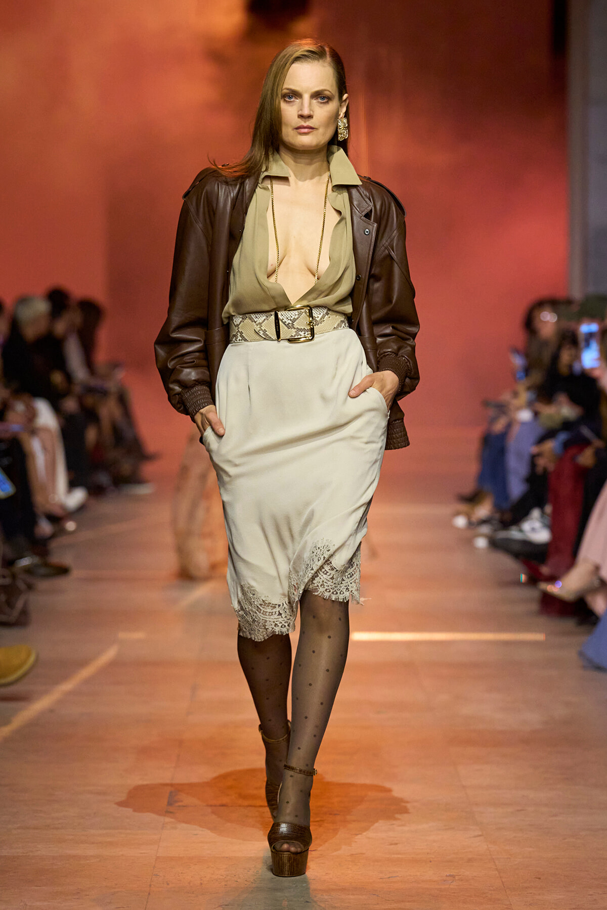 Model walks a runway in a taupe blouse with a plunging neckline, a brown leather jacket, and a cream lace-trimmed skirt with a snakeskin belt.