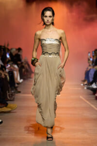 Model on a fashion runway wearing a beige strapless dress with a snakeskin belt and a draped, gathered skirt; orange lighting in background.