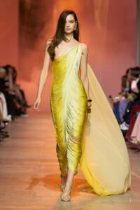 Model walking a fashion runway in a yellow, ruched, one-shoulder gown with a flowing cape trailing behind her.