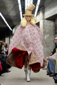 Model walks a runway in an oversized pink crochet cape with red lining, beige hat, and fluffy collar on an industrial-looking set.