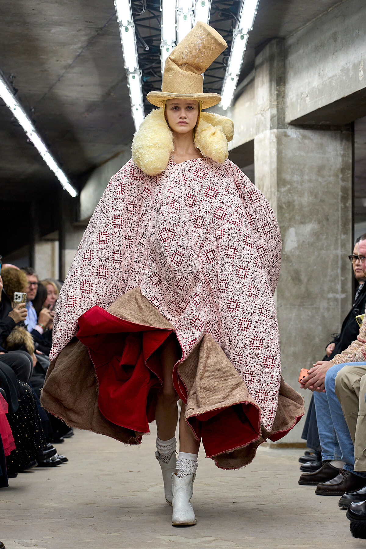 Model walks a runway in an oversized pink crochet cape with red lining, beige hat, and fluffy collar on an industrial-looking set.