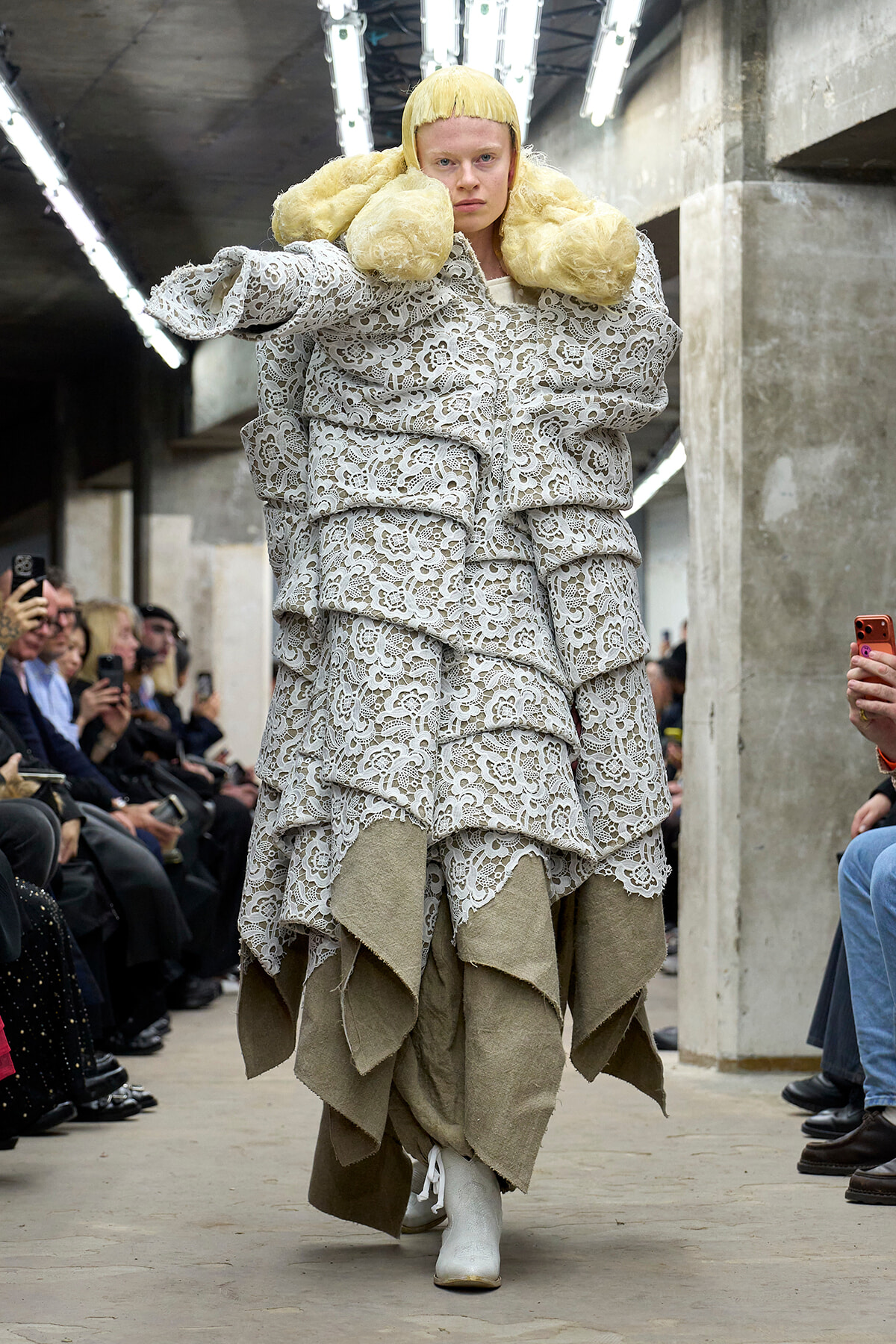 Model walking a fashion runway in an oversized white lace dress with layered ruffles, a pale underlayer, and white boots, wearing a bright yellow wig.