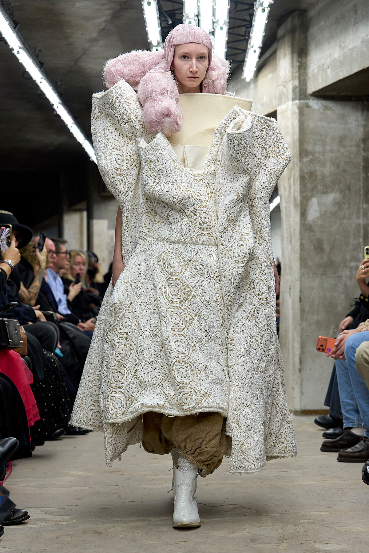 Model on a runway wearing an elaborate white lace gown with oversized sculptural shoulders and pink fluffy hair accessories.