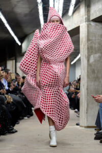 Model on a runway wearing a pink oversized lattice cocoon dress with hood and white ankle boots; audiences line the sides.