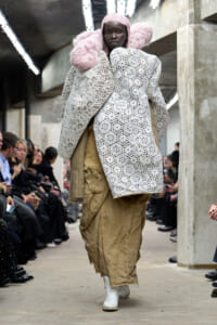 Model walks the runway in a white crochet coat with pink hair and oversized sleeves, paired with rough burlap pants and white boots in an industrial venue.