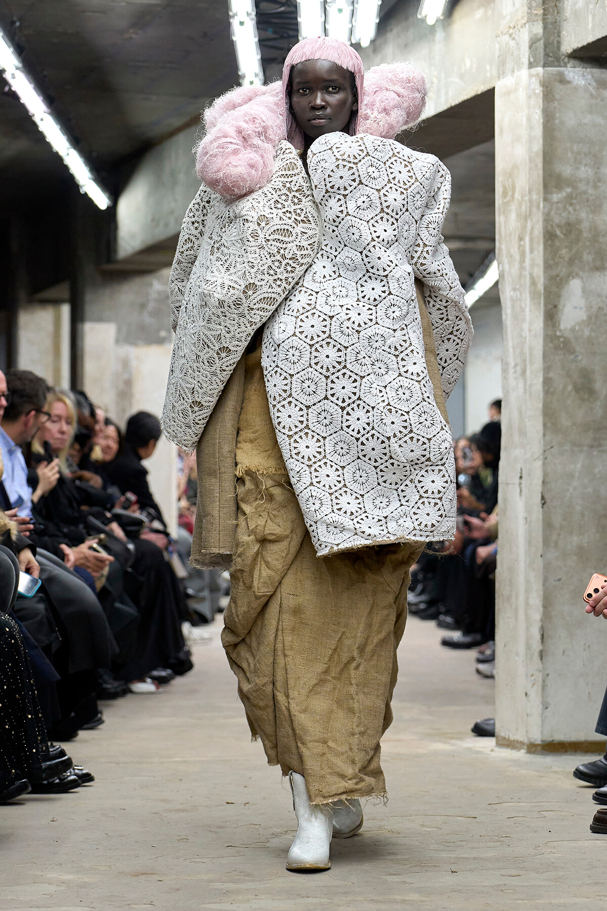 Model walks the runway in a white crochet coat with pink hair and oversized sleeves, paired with rough burlap pants and white boots in an industrial venue.