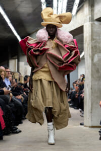 Model walks a runway in a voluminous, beige burlap ensemble with pink ruffle accents, a large beige hat, and white boots; audience watches.