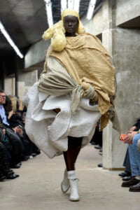 Model walks a fashion runway wearing a layered beige fabric wrap over a white brocade dress, with a yellow wig and white ankle boots.