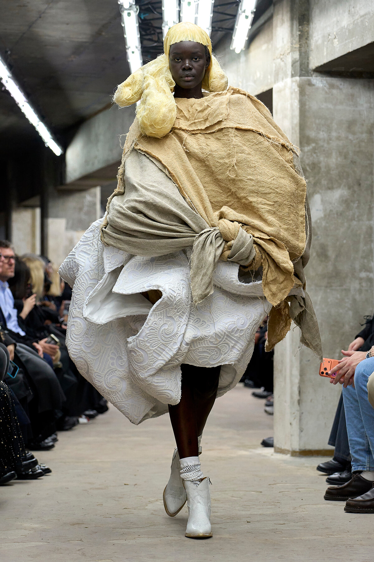 Model walks a fashion runway wearing a layered beige fabric wrap over a white brocade dress, with a yellow wig and white ankle boots.