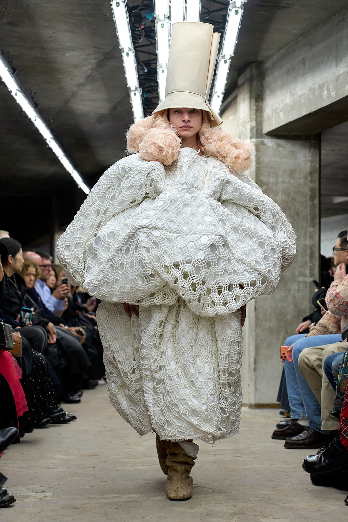 Model wearing a white crochet gown with oversized sleeves and a tall beige hat on a runway runway show scene.
