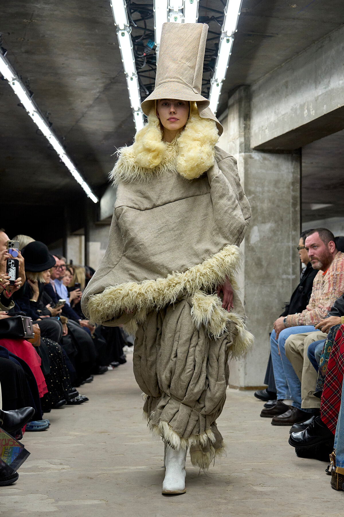 Model walks a fashion runway in a voluminous beige outfit with a tall hat and cream fur trim, surrounded by seated audience and photographers.