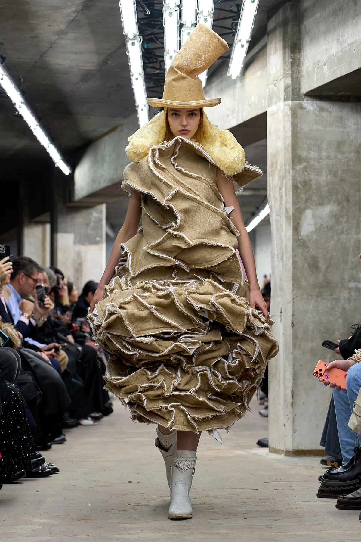 Model walks runway in oversized, sculptural beige burlap gown with layered ruffles and a tall beige hat in an industrial venue with seated audience along the sides