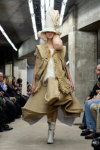Model on a runway wearing oversized beige textured coat with a white fur top and a tall sculptural hat, in an industrial venue with seated audience nearby.