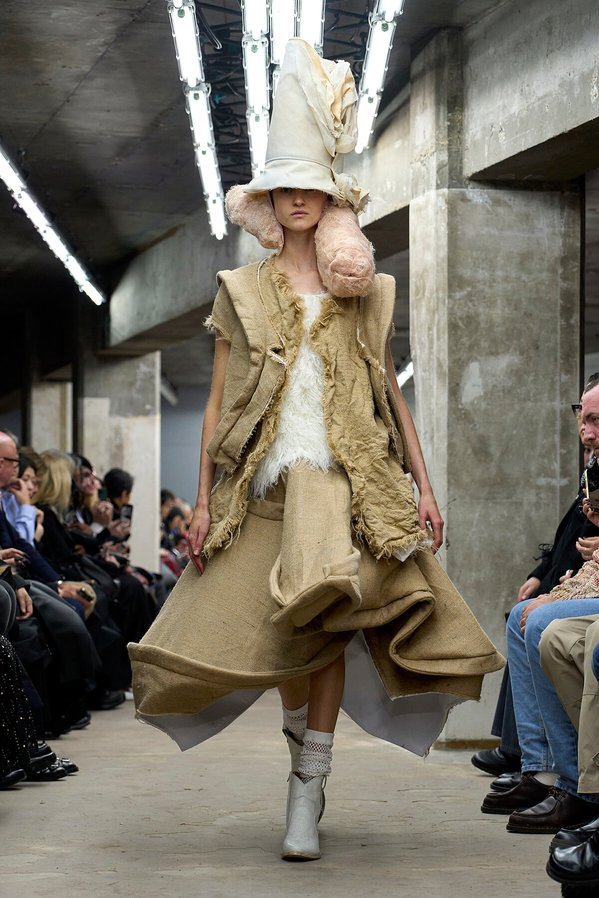 Model on a runway wearing oversized beige textured coat with a white fur top and a tall sculptural hat, in an industrial venue with seated audience nearby.