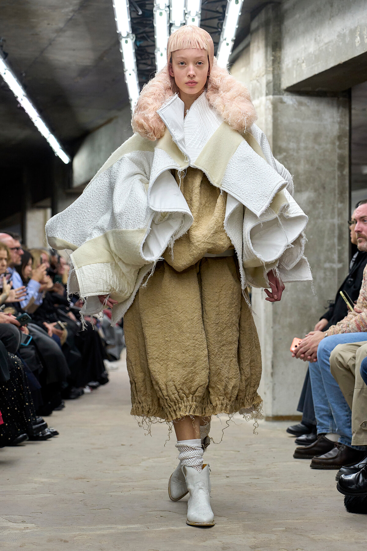 Model on runway wearing an oversized white and cream textured coat with a fluffy pink collar, paired with a tan textured skirt and white ankle boots.