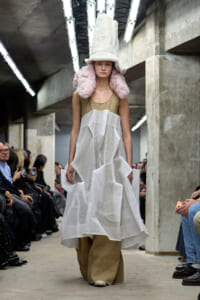 Model walks a fashion show in an industrial venue wearing an oversized white fur hat with pink fluffy ear coverings, a beige top, and a voluminous white mesh skirt over burlap pants, with spectators on both sides.