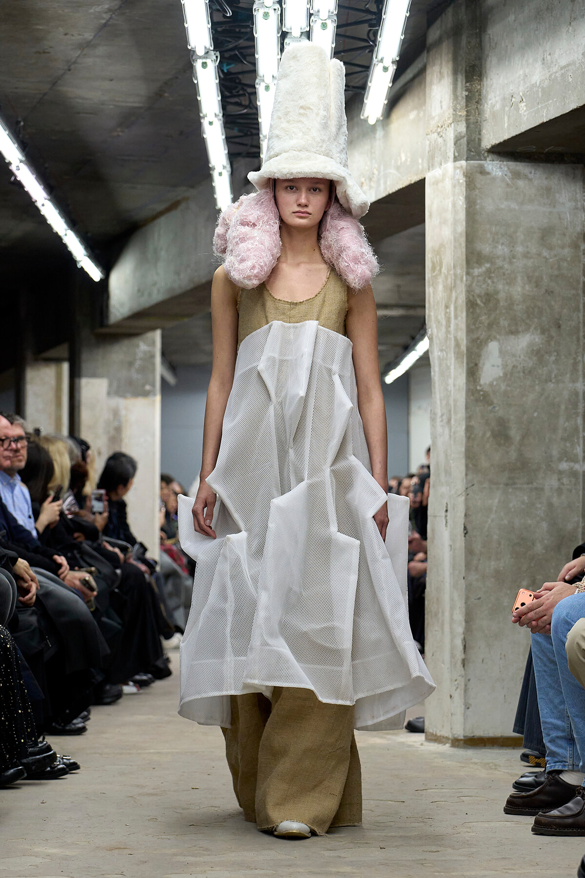 Model walks a fashion show in an industrial venue wearing an oversized white fur hat with pink fluffy ear coverings, a beige top, and a voluminous white mesh skirt over burlap pants, with spectators on both sides.