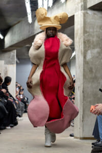 Model on a runway wearing a sculptural beige shell coat with a red interior and oversized hat.