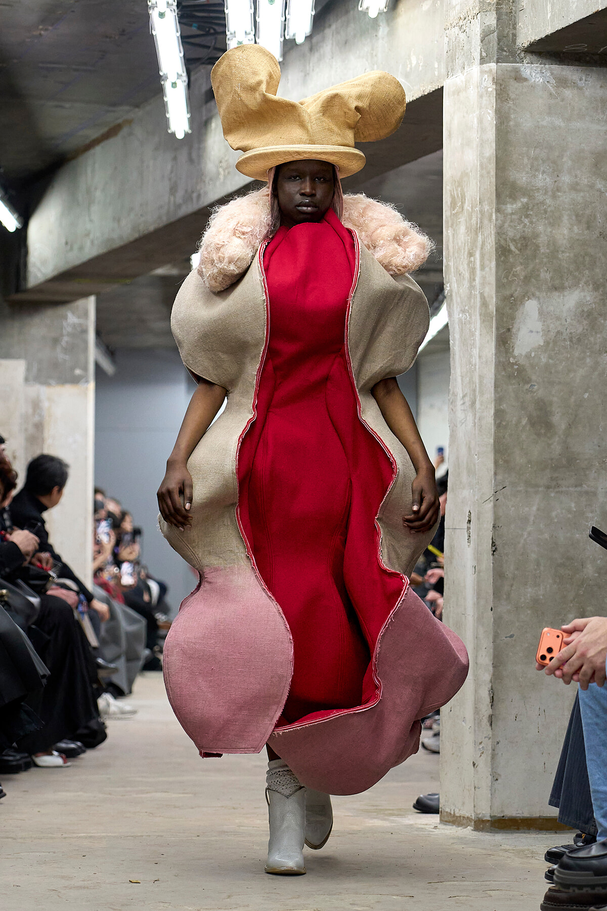 Model on a runway wearing a sculptural beige shell coat with a red interior and oversized hat.