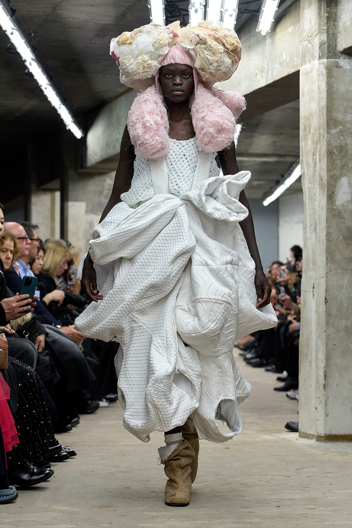 Model walks a runway in a dramatic white textured gown with oversized draped ruffles and pink fluffy neckpieces, in an industrial venue with seated audience.