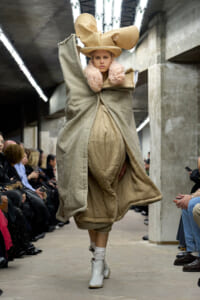 Model walks a runway in an oversized beige ensemble with a large hat and pink faux-fur collar, set in an industrial venue with seated spectators on both sides.