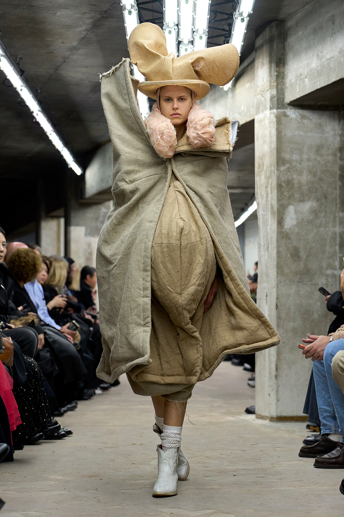 Model walks a runway in an oversized beige ensemble with a large hat and pink faux-fur collar, set in an industrial venue with seated spectators on both sides.