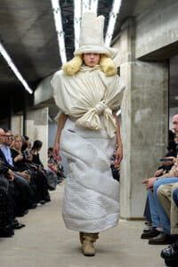 Model walks a fashion show in an oversized cream outfit with a tall sculptural hat and fur accents, industrial runway setting behind.
