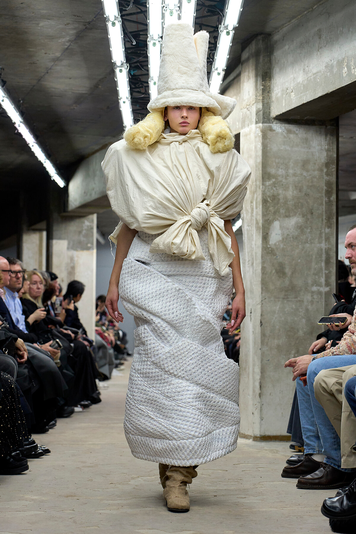 Model walks a fashion show in an oversized cream outfit with a tall sculptural hat and fur accents, industrial runway setting behind.
