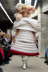 Model on a runway wearing a voluminous white layered dress with a red hem, oversized fabric headpiece, white boots, and seated audience visible along the sides.