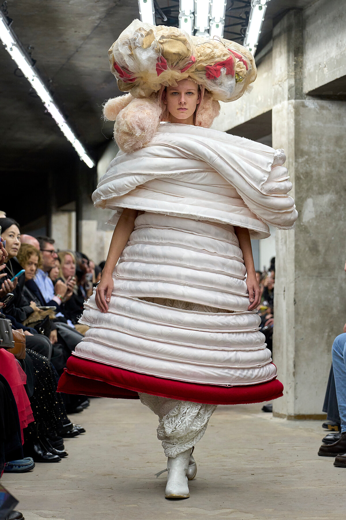 Model on a runway wearing a voluminous white layered dress with a red hem, oversized fabric headpiece, white boots, and seated audience visible along the sides.
