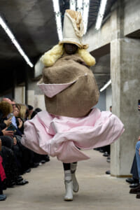 Model walking a runway in a voluminous pink gown with a giant burlap headpiece and tall beige hat, audience seated along a concrete-industrial venue.
