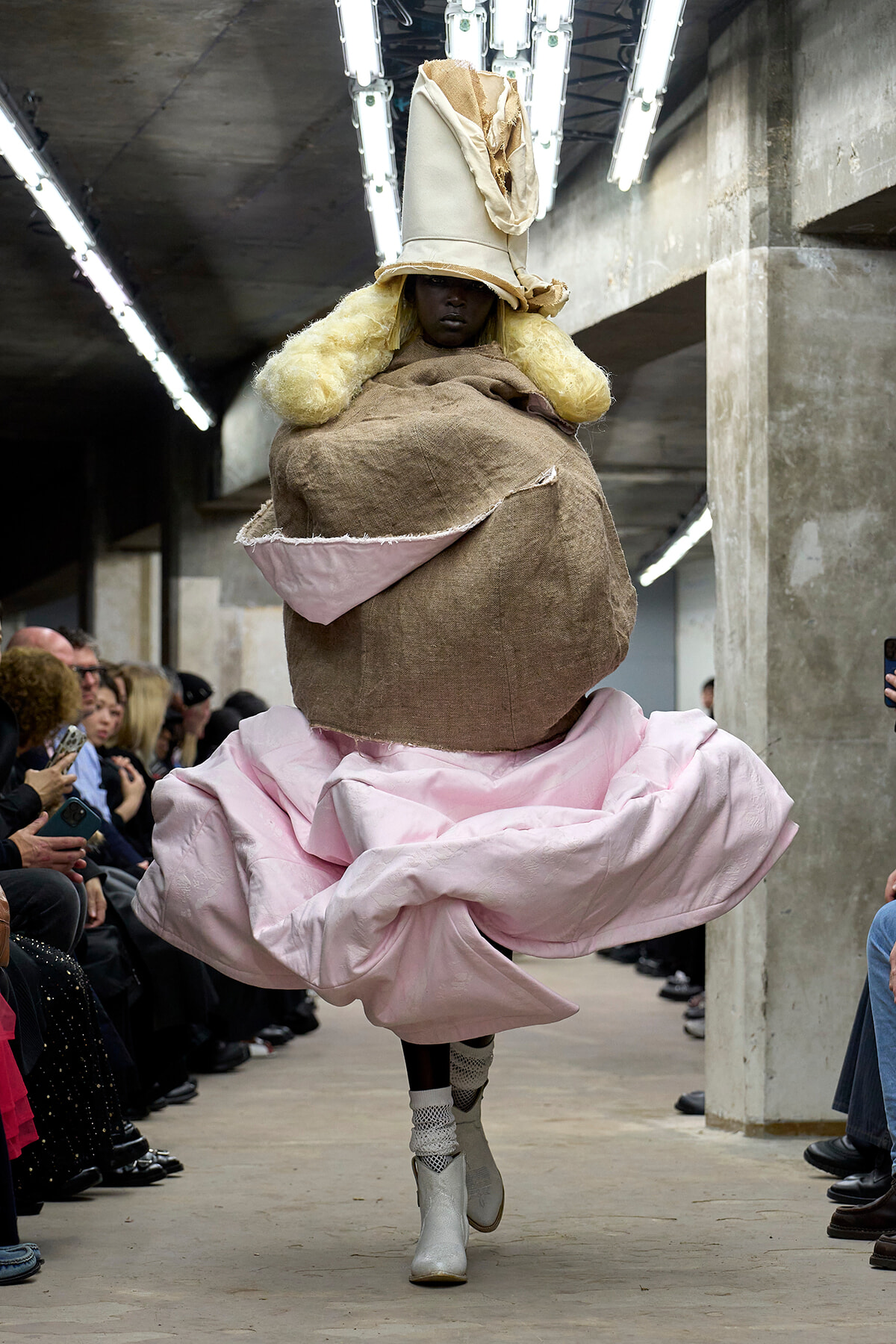 Model walking a runway in a voluminous pink gown with a giant burlap headpiece and tall beige hat, audience seated along a concrete-industrial venue.
