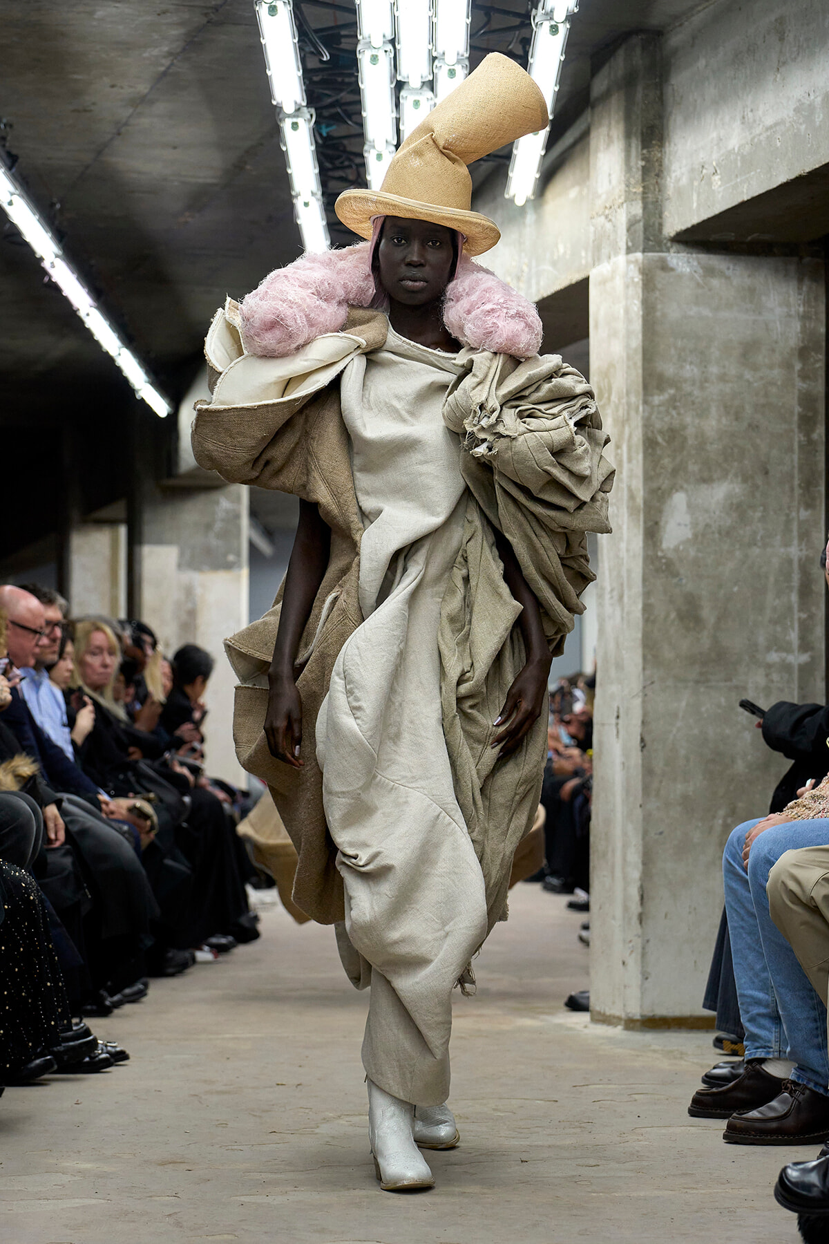 Model walks a fashion runway in a sculptural beige layered gown with pink fur collar and a tall beige hat in an industrial venue.